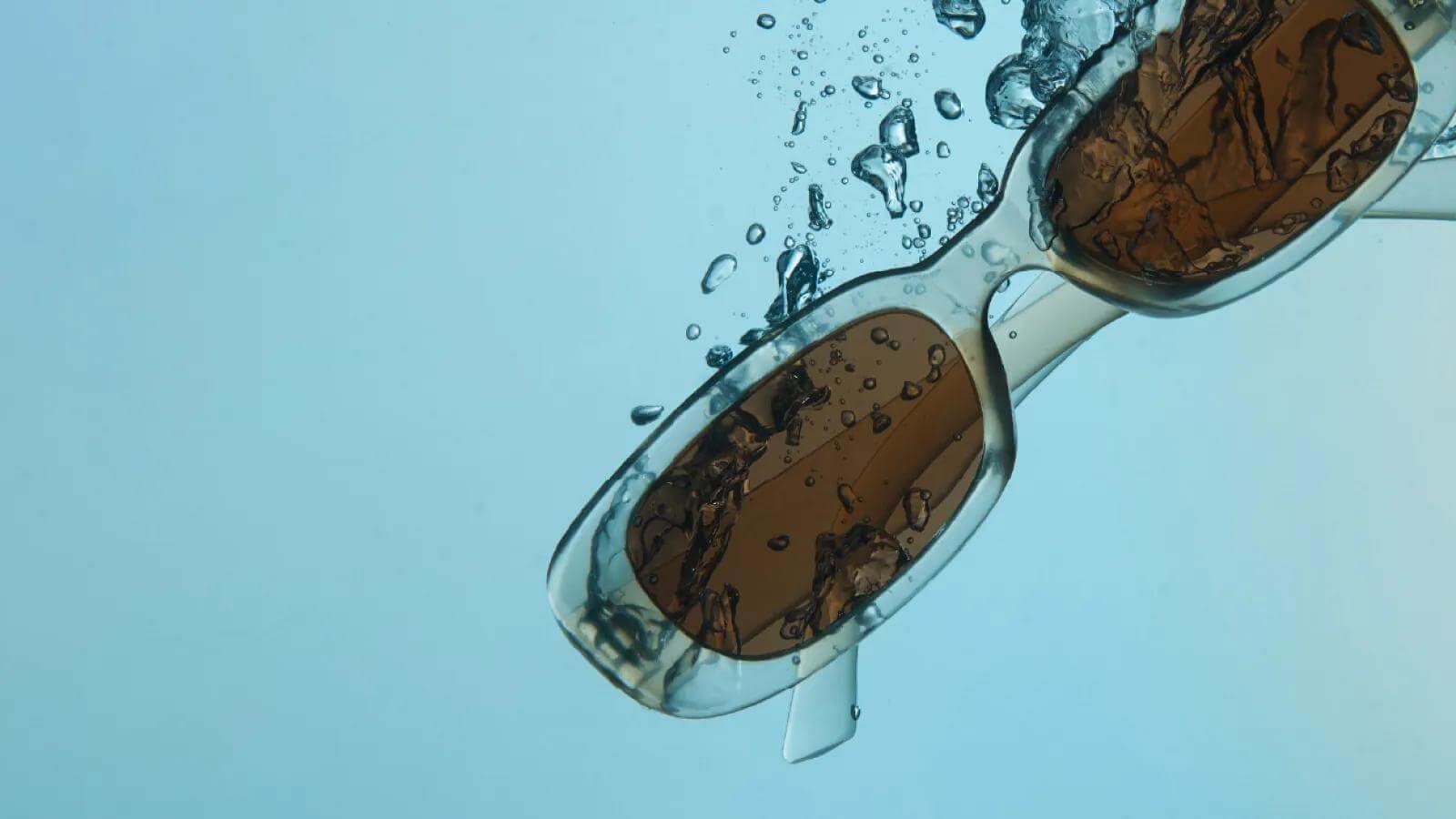 A creative product photo of retro square-frame sunglasses with a translucent icy-blue frame and brown lenses, surrounded by splashing water against a soft sky-blue background, perfect for eyewear marketing.